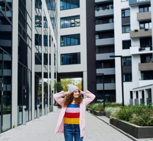 Front view portrait of young woman standing outdoors by block of flats in city.